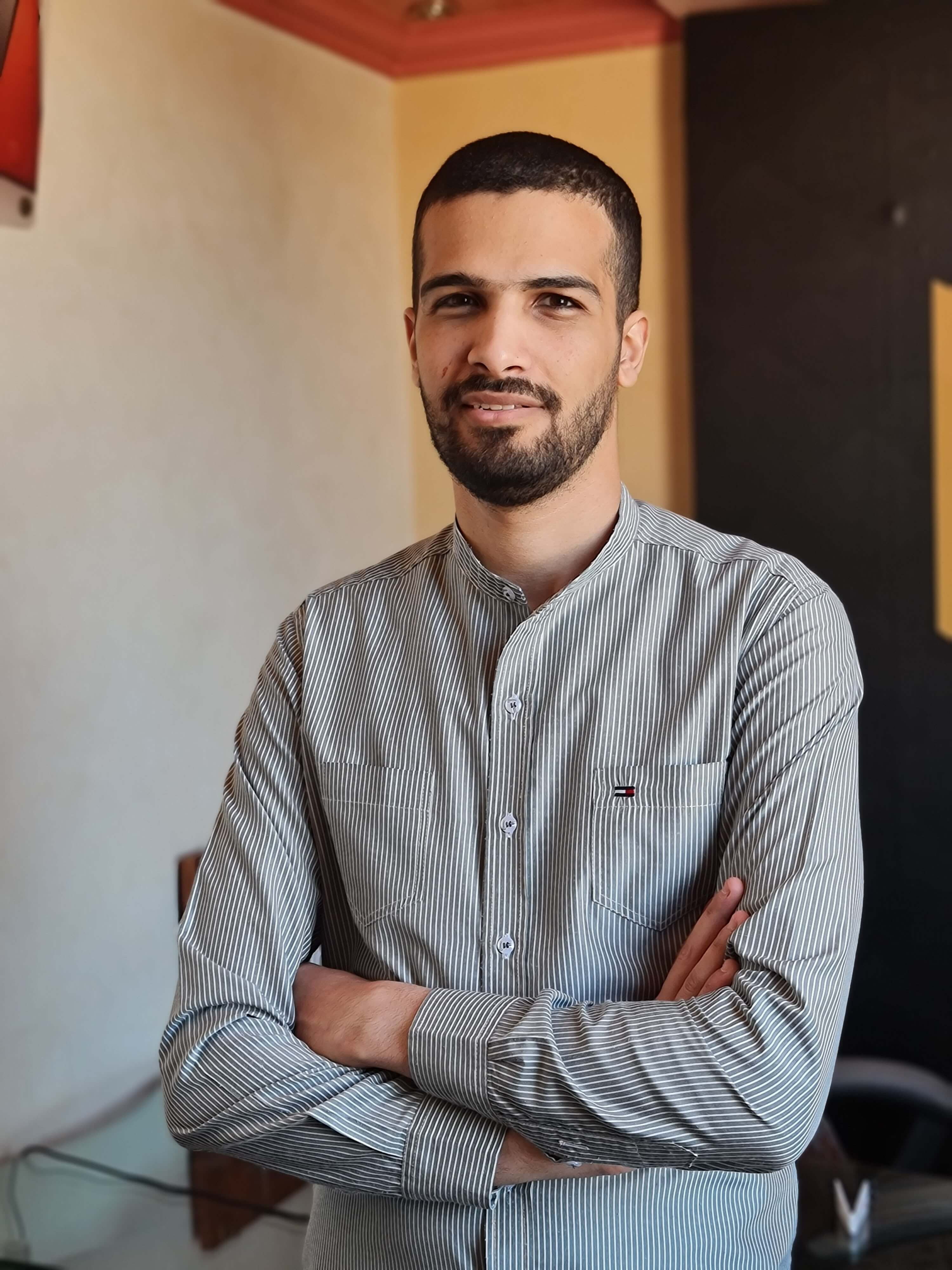 Mohamed Tanash standing indoors with his arms crossed. He has short dark hair, a trimmed beard, and is wearing a light-colored, vertically striped shirt with buttons. The background features a neutral wall and some subtle elements of furniture and decor, such as a chair and a corner with contrasting colors. The lighting is soft and natural.